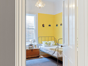 Yellow feature wall corner in bedroom with white bedding and wooden side table with photos carpet